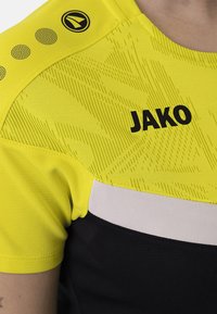 Maglietta sportiva a maniche corte con parte superiore gialla neon caratterizzata da un motivo testurizzato, sezione inferiore nera e un grande logo "JAKO" in nero.