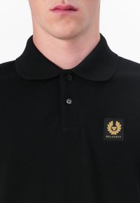 Must värvi polo särk klassikalise kraega, kolme nööbiga rinnaehtes, struktureeritud kangast ja vasakul rinnal kuldne logo märgis.