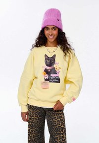 Žlutá mikina s grafikou černé kočky a květin, v kombinaci s kalhotami s leopardím vzorem a růžovou pletenou čepicí.