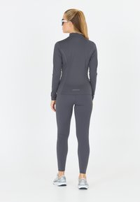 Grå langærmet atletisk top med tætsiddende design, parret med matchende leggings. Modellen bærer reflekterende solbriller og løbesko.