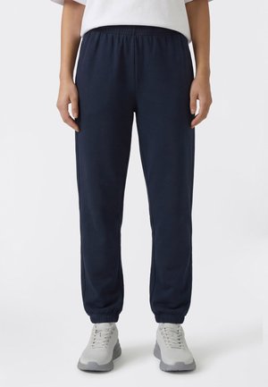 Osoba ubrana w granatowe spodnie dresowe z elastycznym pasem i białe tenisówki, stojąca na tle jasnego, jednolitego tła.