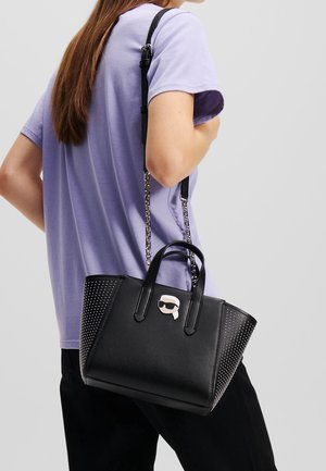 Borsa nera con lati perforati e un emblema decorativo. Presenta doppi manici e una tracolla rimovibile con catene.