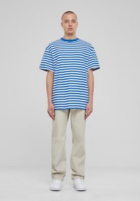 Mees seisab seljas sinise ja valge triibulise lühikese varrukaga särk, beežid püksid ja valged spordijalatsid, mille taustal on sile hallivalge taust.