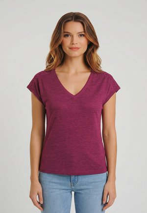 Młoda kobieta o falowanych włosach, ubrana w fioletową koszulkę z krótkim rękawem i dekoltem w literę V oraz jasnoniebieskie dżinsy, stoi na tle jednolitego jasnego tła.