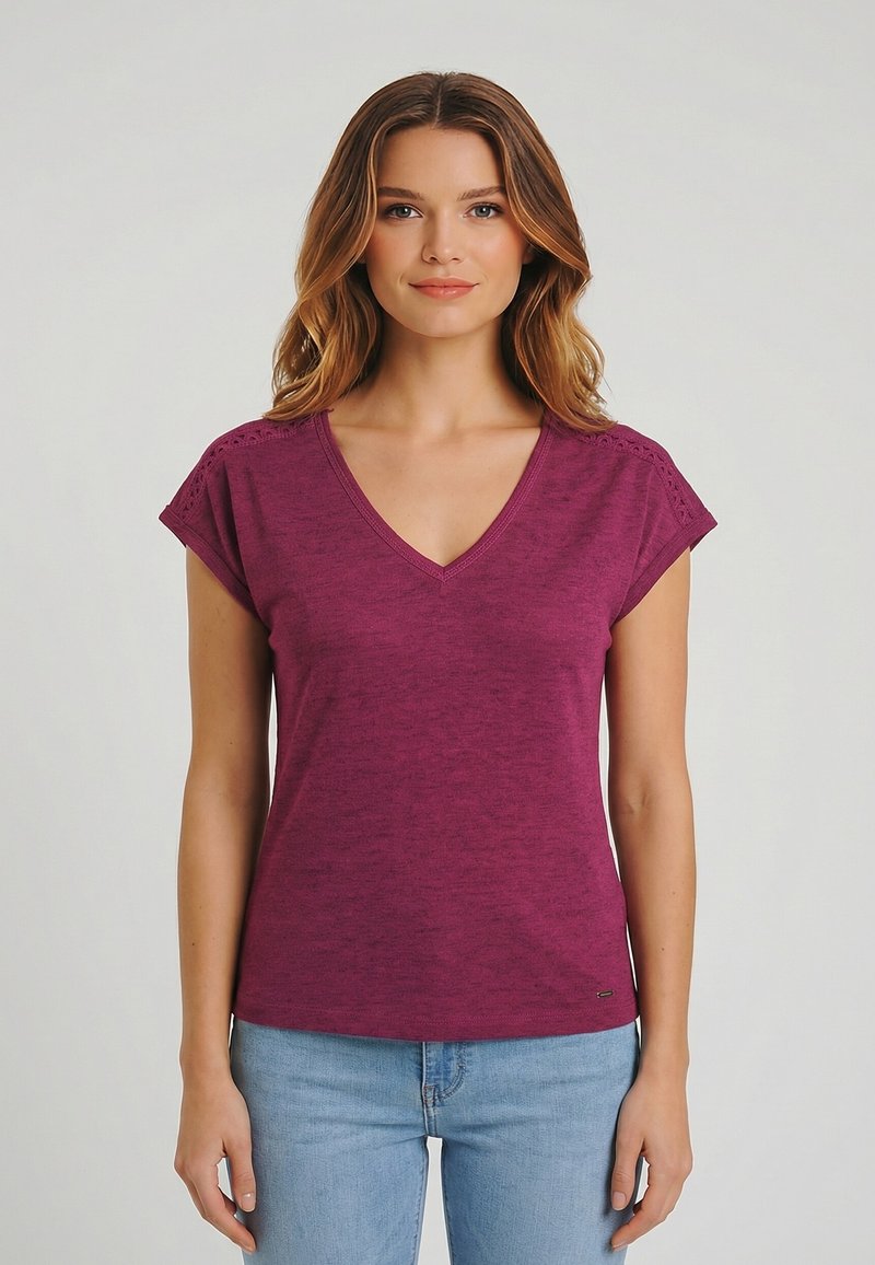 Giovane donna con capelli mossi che indossa una maglietta viola a maniche corte con scollo a V e jeans azzurri, in piedi contro uno sfondo chiaro e uniforme.