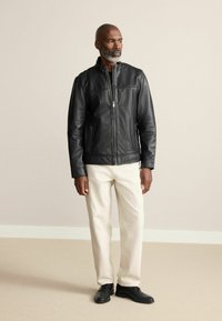 Schwarze Lederjacke mit einem Frontreißverschluss, Stehkragen und zwei seitlichen Taschen. Trägt man über hellen Hosen und schwarzen Schuhen.