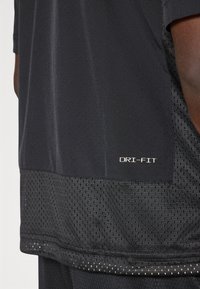 Fekete DRI-FIT sportpóló, félig áttetsző, légáteresztő textúrával, sima felső résszel és hálós alsó panellel.