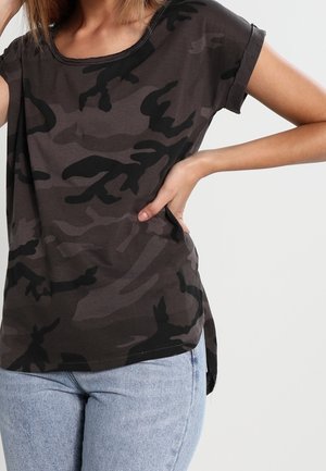 Woman wearing a dark gray and black camouflage short-sleeve shirt and light blue jeans, with one hand resting on her hip.