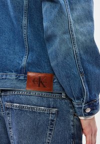 Μπλε τζιν μπουφάν με δερμάτινο ταμπελάκι με την ένδειξη "Calvin Klein" στην πλάτη, διαθέτει εφαρμοστή γραμμή, μανσέτες με κουμπιά και εμφανείς ραφές.