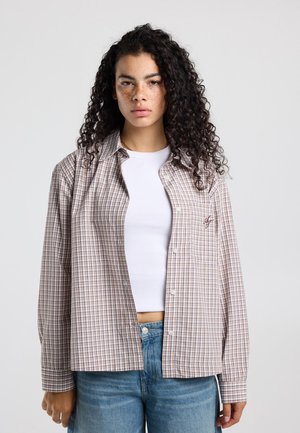 Vrouw met krullend haar die een geruit hemd draagt over een wit crop top en een blauwe spijkerbroek, staand tegen een effen achtergrond.