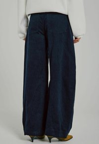 Weite Cord-Hosen in dunkelblau mit vertikaler Ripptextur, hoher Taille und zwei Gesäßtaschen. Kombiniert mit braunen Absätzen.