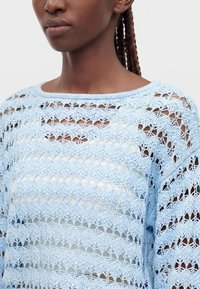Femme aux cheveux tressés portant un pull bleu clair à mailles ouvertes par-dessus un top blanc, visible des épaules jusqu'au milieu de la poitrine sur un fond uni.