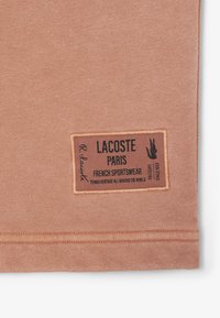 Tela de color melocotón con una textura suave y una etiqueta rectangular con texto negro y un logo, que dice "Lacoste Paris French Sportswear."