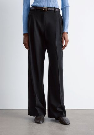 Person wearing black wide-leg trousers with a brown belt, blue ribbed long-sleeve top, and dark brown shoes standing on a gray floor.