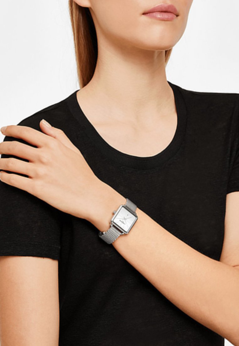 Zilveren rechthoekige klok met een mesh band om de pols. Heeft een witte wijzerplaat met subtiele markeringen en een minimalistisch ontwerp, gecombineerd met een zwart shirt.