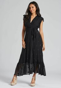Robe noire mi-longue avec des manches volantées, ceinture à nouer, motifs en diamants brodés et couche inférieure transparente. Associée à des sandales compensées.