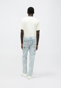 Camiseta de color beige claro, corte relajado, combinada con jeans azul claro que tienen varios bolsillos y zapatillas blancas. Fondo neutro.