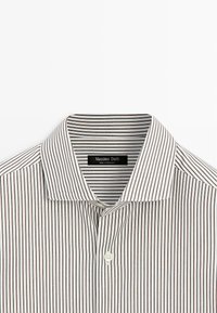 Chemise rayée avec un col pointu, présentant des rayures verticales alternées blanches et marron, et une fermeture à un bouton au niveau du cou.