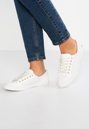 Personne portant un jean bleu slim et des baskets basses blanches avec des lacets blancs, debout sur un sol lisse de couleur claire.