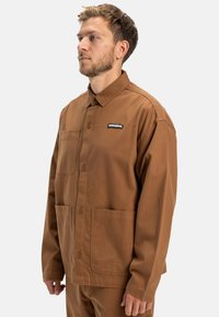 Braune Arbeitsjacke aus robustem Stoff, mit knöpfbarem Frontverschluss, einer Brusttasche und zwei unteren Taschen. Sichtbares Logo-Label auf der Brust.