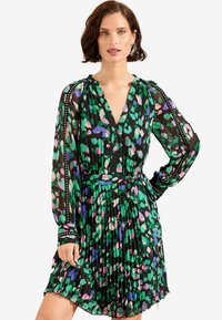 Rochie cu imprimeu floral, cu accente verzi, mov și roz pe un fundal negru. Are decolteu în V, mâneci lungi și fustă plisată.