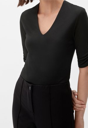 Kobieta ubrana w dopasowaną czarną bluzkę z dekoltem w serek i rękawami do łokcia oraz wysokim stanem czarne spodnie, trzymająca lewą rękę za plecami.