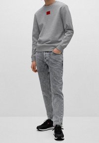 Grauer Pullover mit kleinem rotem Etikett, kombiniert mit hellgrauen Jeans und schwarzen Turnschuhen. Schlichtes Design, weiche Textur, legere Passform.