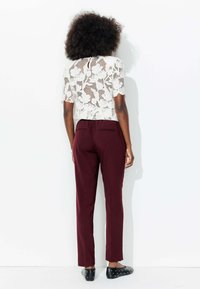 Haut en dentelle à motif floral en blanc cassé, associé à un pantalon bordeaux ajusté. Chaussures noires avec des accents cloutés. Vue arrière, fond minimal.