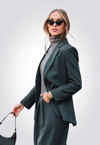 Donkergroen op maat gemaakte blazer over een grijze coltrui, gecombineerd met bijpassende broek. Accessoires omvatten gouden armbanden en een zwarte handtas.