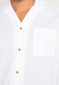 Camicia button-up bianca in lino con taschino sul petto, texture morbida e bottoni arancioni. Collo corto con vestibilità comoda.
