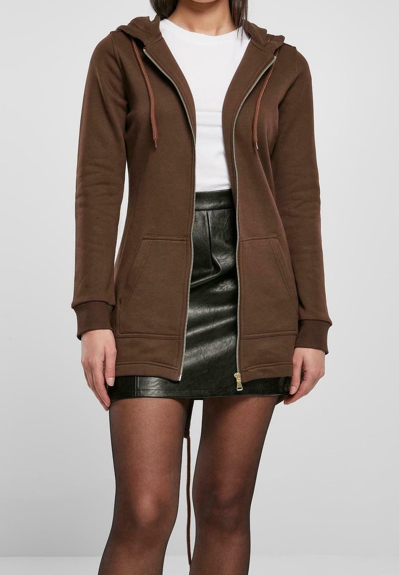 Femme portant un sweat à capuche marron zippé, un haut blanc, une mini-jupe en cuir noir et des collants noirs transparents, debout devant un fond gris clair.