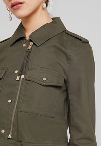 Zbliżenie oliwkowej kurtki z zamkiem błyskawicznym z przodu, kieszeniami na zatrzaski, naramiennikami, noszonej przez osobę z wiszącymi perłowymi kolczykami.