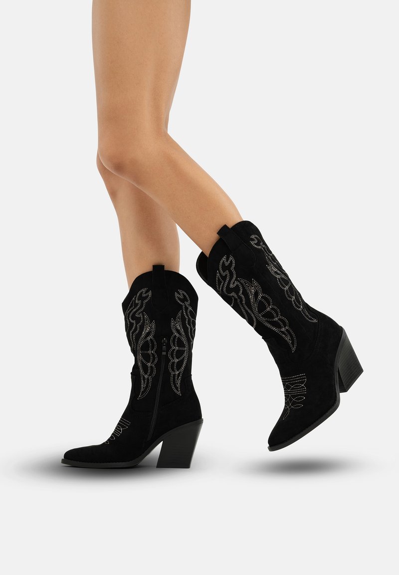 Botas de cowboy de ante negro con bordado curvado en los lados, punta cuadrada y un tacón de altura media y acampanado. Presentan una cremallera en el lado.