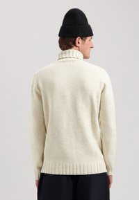 Cremfarbener Strickpullover mit hohem, geripptem Kragen. Der Pullover weist ein strukturiertes Muster sowie Rippen am Saum und an den Bündchen auf.