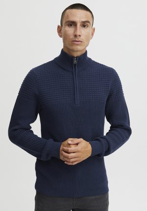 Pulover tricotat de culoare albastru marin, cu guler înalt, fermoar frontal, corp texturat și manșete și tiv cu nervuri. Prezentând o combinație de modele.