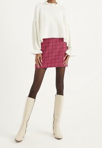 Vit kort topp med volangkrage och puffärmar, i kombination med en röd rutig minikjol. Beige knähöga klackstövlar med dragkedjor.