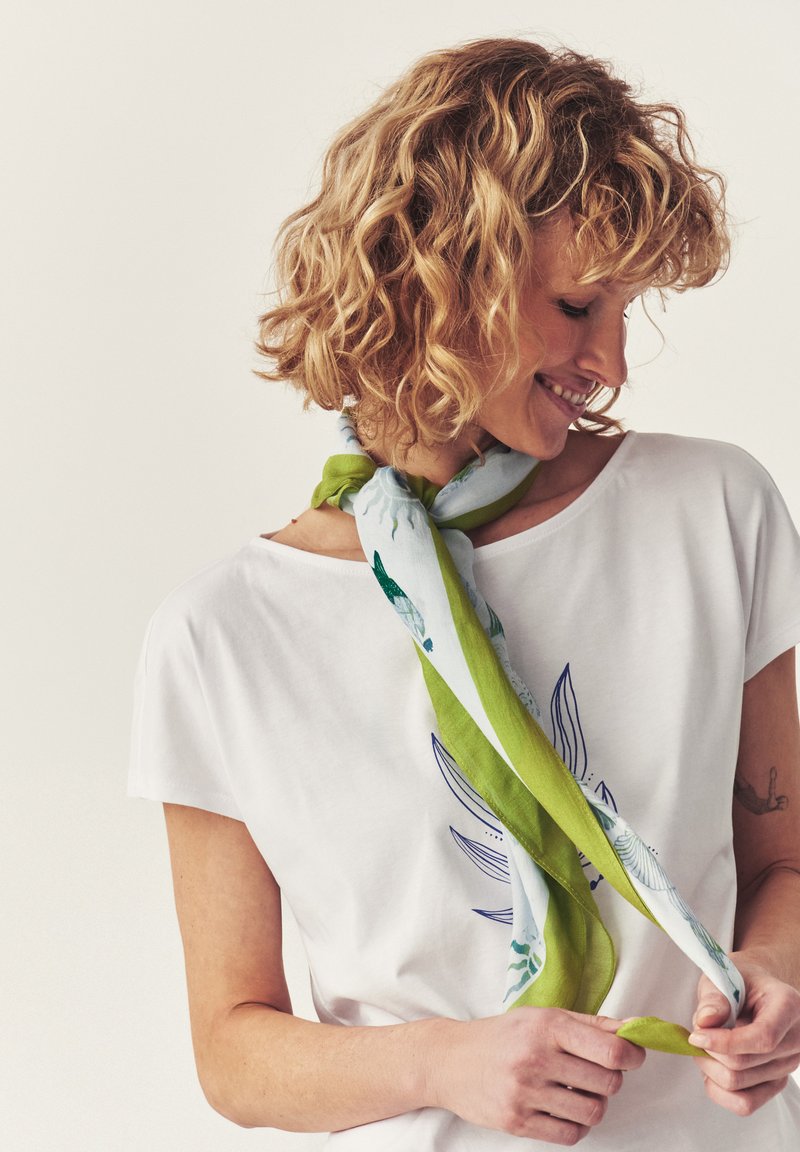 Femme aux cheveux blonds bouclés souriante, portant un t-shirt blanc et nouant un foulard vert et blanc à motifs autour du cou.