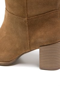 Gros plan d’une bottine en daim marron avec un talon bloc en bois empilé et des détails de couture visibles.