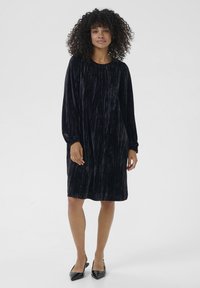 Robe en velours noir à manches longues, coupe décontractée et texture verticale. Le mannequin porte des escarpins noirs pointus, debout avec les bras détendus.