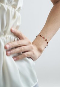 Brățară din aur cu cristale roșii în formă de lacrimă, accente circulare și un finisaj neted și detaliat, purtată pe încheietura mâinii.