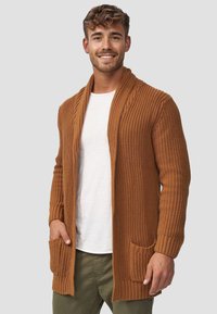 Lang, strikket cardigan i en varm brun farve med et ribbet mønster. Har en sjalskrave og to forlommer. Båret med en hvid skjorte.