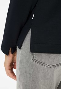 Zwart shirt met lange mouwen en zijsplitten, gedragen over lichtgrijze spijkerbroek. Zichtbare stiknaden en een klein label aan de zijkant van het shirt.