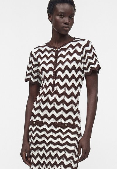 Femme portant une robe marron et blanche à motif zigzag, manches courtes, détails à boutons sur le devant et poches.