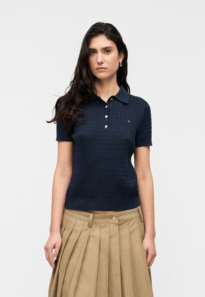 Ženska z dolgimi temnimi lasmi, oblečena v mornarsko modro kratkoročno pleteno polo majico z gumbi in bež zloženo krilo, stoji pred belim ozadjem.