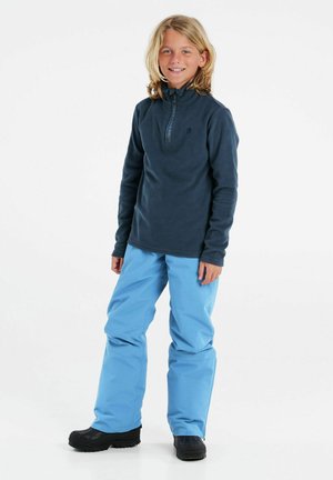Pullover in pile blu navy con colletto alto e zip a quarto, abbinato a pantaloni impermeabili azzurri chiari e scarpe nere.