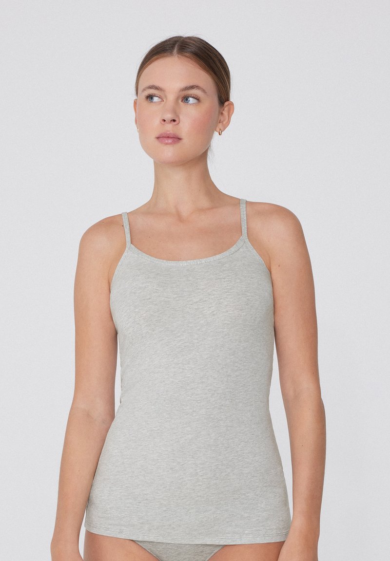 Frau trägt ein hellgraues ärmelloses Tanktop mit dünnen Trägern und blickt seitlich vor einem einfachen weißen Hintergrund.