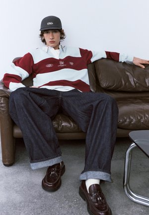 Jeune personne en maillot de rugby rayé, jean large foncé, mocassins à pampilles et casquette, allongée sur un canapé en cuir marron dans une pièce moderne.