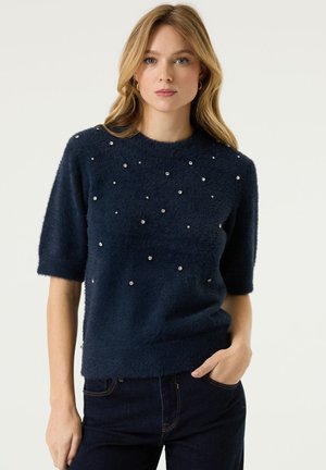 Vrouw staat met één hand in de zak, draagt een navyblauwe, pluizige trui met korte mouwen en kleine parelachtige versieringen, en een donkere spijkerbroek.
