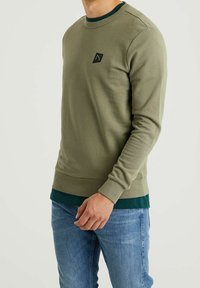 Olivgrön sweatshirt med ribbad rundhals och mudd i ärmsluten. Har en liten svart logotypklistermärke på bröstet. Bärs över en mörkgrön skjorta.