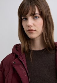 Donkerrode pufferjas met capuchon en merkdetail, gedragen over een donkerbruine gebreide trui. Gladde textuur en een casual pasvorm.